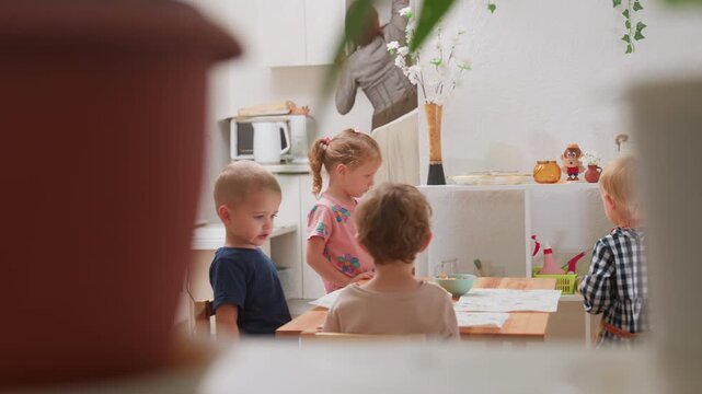 Children engaged around table while instructor reaches shelf and hands book to girl in pink clothing during kindergarten activity with classmates observing showing cooperation learning responsibility