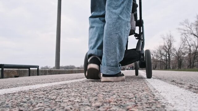 A person wearing jeans and sneakers pushes a baby stroller along an asphalt path. The ground-level perspective captures the rhythm of the walking feet and rolling wheels.