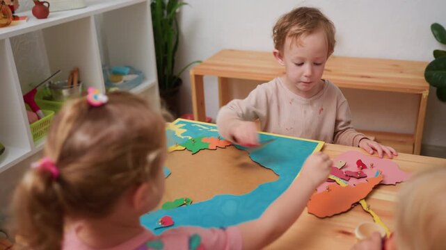 preschool kids move continent puzzle pieces from map board to side, practicing geography and fine motor skills in bright montessori classroom, wooden tables, shelves, plants, collaborative learning