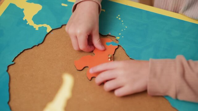 student hands position orange continent puzzle piece into map board, guiding knob into outline on turquoise water, careful alignment for geography lesson, fine motor control