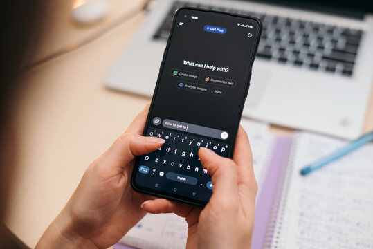 Kyiv, Ukraine - October 12, 2025: Person typing on smartphone using AI chat GPT app, with blurred notebook and laptop in background, modern workspace concept.