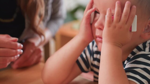 Autistic child carrying picked block and placing it on cheek while woman watches closely, striped shirt, colorful blocks on wooden table, attentive supportive guidance in inclusive classroom