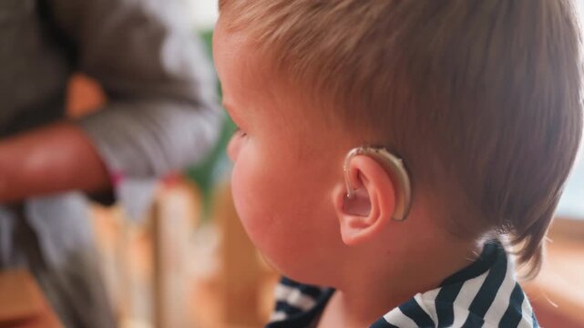 Mother guides her special child with hearing aid during supportive moment indoors, close side view highlighting ear device and striped shirt, nurturing guidance in warm environment