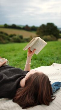 A woman is laying on a blanket reading a book. The scene is peaceful and relaxing, with the woman enjoying her time outdoors