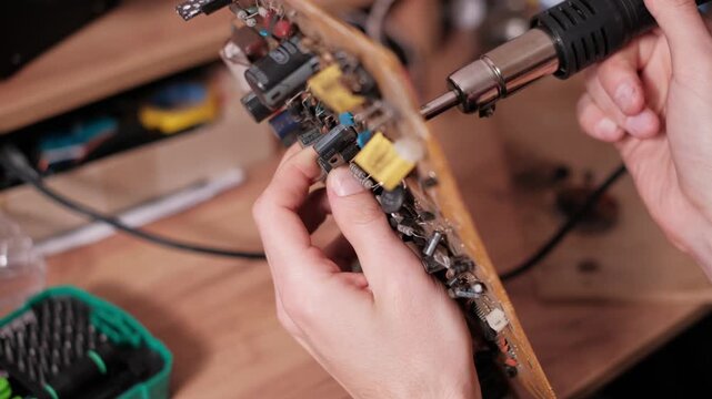 Hands carefully position a hot air gun nozzle to heat and desolder a specific component from the front side of a printed circuit board during a detailed electronics repair or rework task..