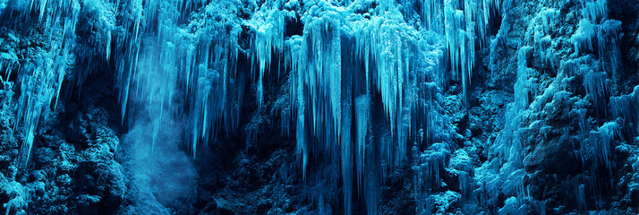 Stalactites of ice dangle from the roof of a cave, reflecting cold blue tones. The ground is rocky, creating a chilly winter scene filled with natural beauty, banner
