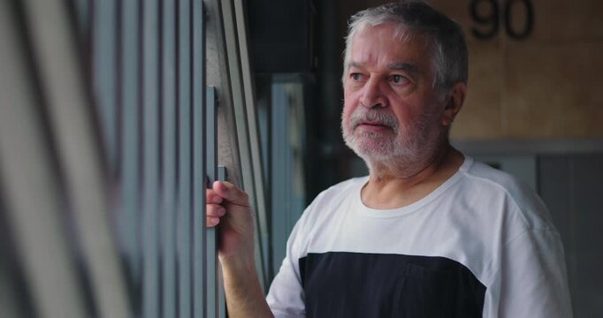 Elderly man standing near a metal gate, holding the bars with one hand, looking outward with a worried expression, symbolizing isolation, surveillance, and introspection