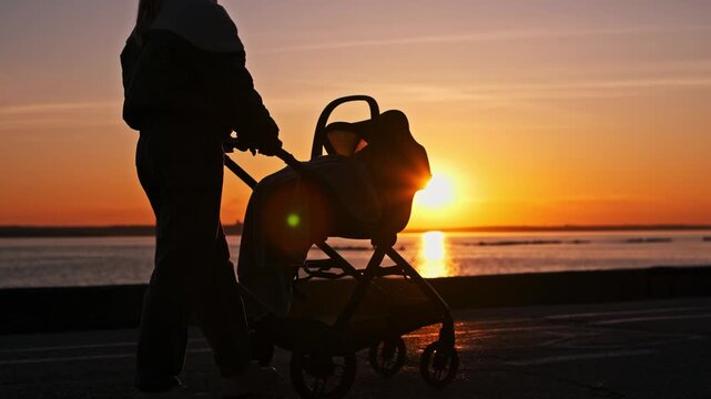 Silhouette of a mother pushing a baby stroller along a waterfront path during an orange sunset, with sun reflecting on calm water, creating a serene evening scene with parent and child.