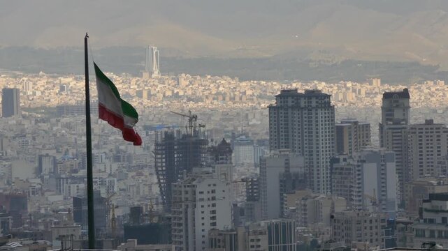 The Iranian flag waves on a tall pole above Tehran skyline, with high-rise buildings, cranes, and mountains in the distance, symbolizing national identity, resilience, and urban transformation.
