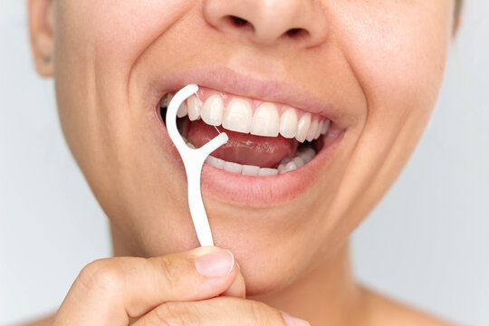 Close-up of a young woman using a dental floss pick to clean between her teeth on a light background. Concept of oral hygiene, fresh breath, and daily dental care routine