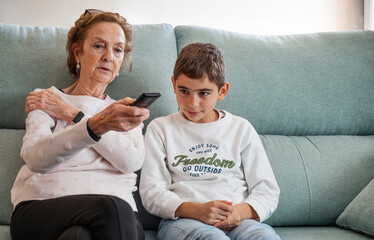 Grandmother and grandson sitting on couch watching TV together at home, intergenerational bonding, quiet family moment with love, learning, togetherness, warmth, childhood and elderly connection