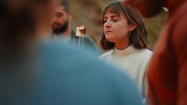 A woman is eating a marshmallow over a fire. The scene is outdoors and the people are gathered around the fire