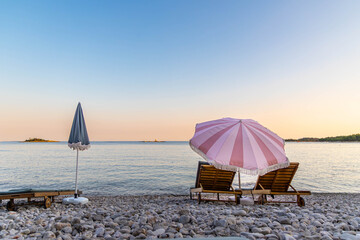 Romantic beach sunset with umbrellas and loungers. Peaceful pebbled beach at sunset with two wooden loungers under a pink umbrella, perfect for romantic or relaxing vacation themes. © Iryna