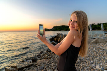 Happy girl taking smartphone photo at sunset beach. Smiling girl captures a scenic sunset on her phone while standing on a rocky beach, enjoying summer vacation by the sea.