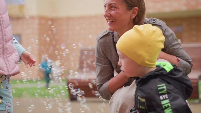 Happy mother bends close watching her son in yellow cap joyfully playing with floating soap bubbles on sunny courtyard, sharing laughter and bonding during cheerful outdoor activity