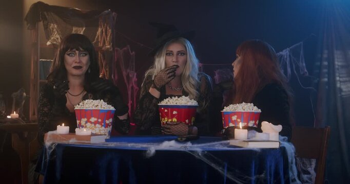 Three women dressed as witches are eating popcorn, watching a movie on TV, and are very excited in a dimly lit, festive Halloween setting with candles. Halloween concept, scary atmosphere, witch party