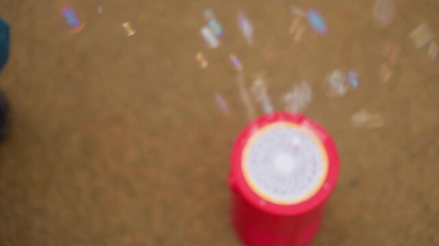 Top view close focus on red bubble machine on pavement releasing streams of soap bubbles upward, circular grille visible, soft blur background enhances shimmering motion