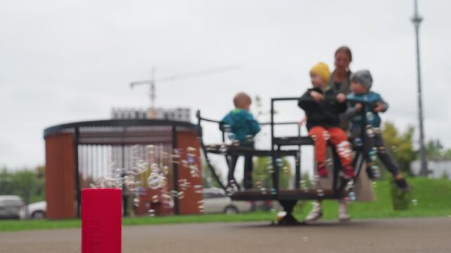 Blur background shows caregiver rotating children on merry go round anticlockwise while red bubble machine blows soap bubbles in foreground, cloudy sky over park, playful spin