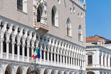 Fototapeta premium Doges Palace Facade with Italian and EU Flags. Close-up of the ornate Gothic facade of Doges Palace in Venice, featuring arched windows, marble columns, and national flags.
