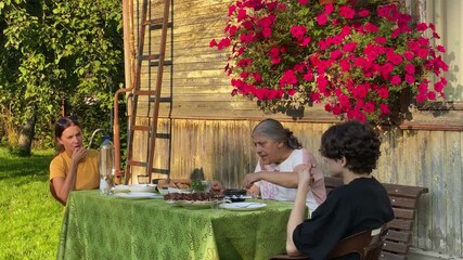 Three generations of women are enjoying an outdoor meal together on a sunny day. They are sitting at a table with food, engaged in conversation and laughter, creating a warm family moment.