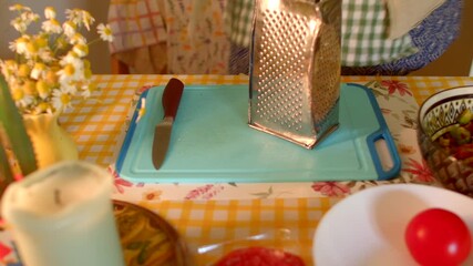 This demonstration showcases skillful grating of fresh vegetables on a blue cutting board, using a metal grater and knife, all against a cheerful yellow checkered tablecloth