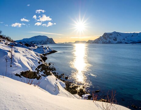 A serene winter landscape of a snow-covered coastline with mountains under a bright sun setting over the water