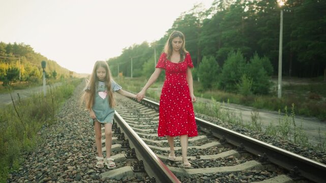 young girl walks on railway sleeper holding mother hand for balance as they move together along countryside track under soft sunlight surrounded by trees and rural path