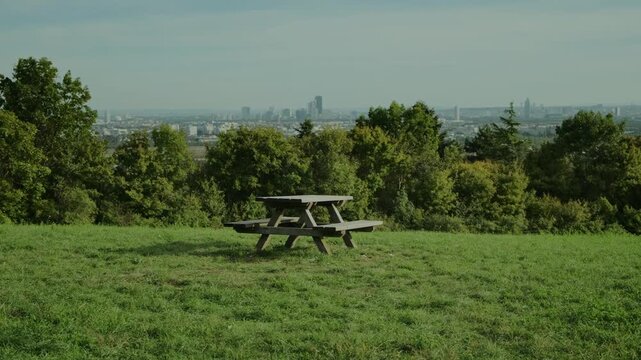 Wooden bench and table on a hilltop with Vienna in the distance. Gentle wind sways the forest behind, creating a lively, serene, and atmospheric outdoor scene under clear sunlight.