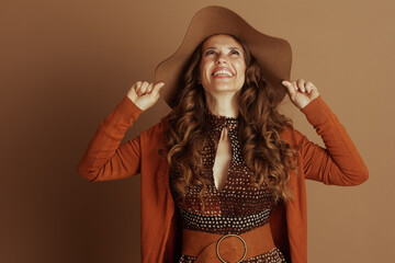 A happy woman in a brown floppy hat and orange cardigan smiles warmly, looking up while gently...