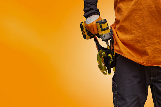 Professional construction worker holding a cordless electric drill with a tool belt full of instruments on bright orange background. Concept of repair, building, maintenance and labor industry.