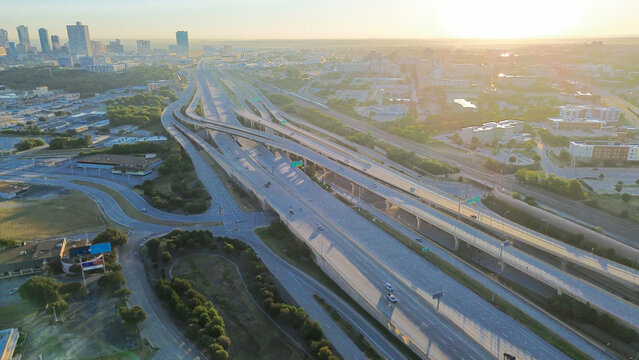 Curved stacked interchange channels traffic into Fort Worth downtown skyline, foreground has logistics hub, flat-roof warehouses, and directional ramps framed by early light and tree buffers