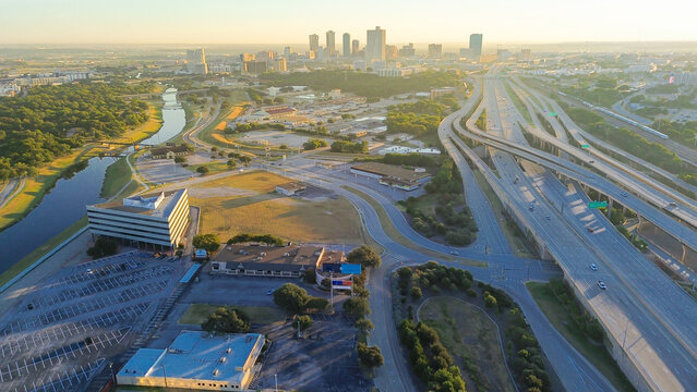 Stacked interchange near Fort Worth divides downtown skyline from industrial logistics zone, elevated ramps, freight access, and golden hour light casting long shadows across both sectors, Texas