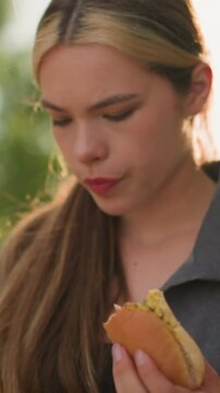Close-up of lady wearing grey top holding shawarma, observing it thoughtfully, soft sunlight illuminates her hand and snack, with a blurred background of greenery and warm sunrays