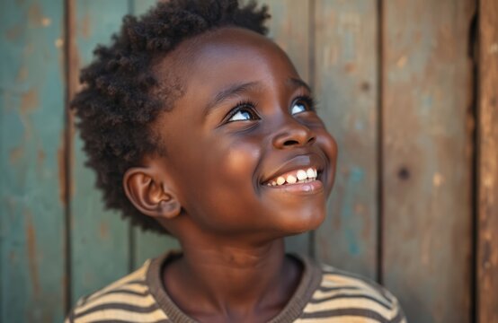 Cute African boy smiling, looking up. Happy child portrait with black skin, short curly hair. Young kid standing near wooden wall. Joyful little boy with striped shirt. African child face close-up. - Powered by Adobe