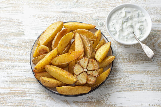 Golden baked potato wedges with garlic, spices and Greek tzatziki sauceon rustic plate. Tasty homemade snack or side dish, healthy vegetarian food
