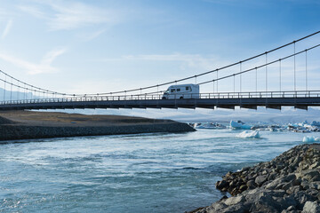 Vehicle crossing a modern suspension bridge over a serene river, surrounded by icy landscapes and clear blue skies, showcasing adventure and exploration in nature