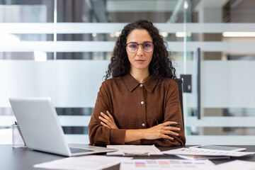 Confident businesswoman in a brown shirt and glasses sits at her office desk with arms crossed,...