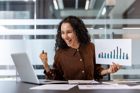 Happy businesswoman at office desk celebrating success, smiling at laptop while holding a financial growth chart and documents during a confident presentation of results