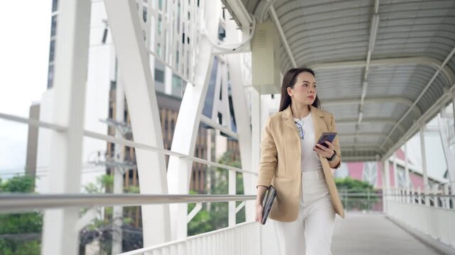 Confident Asian businesswoman walks on urban skywalk holding tablet. Concept of modern leadership, confidence, and ambition.