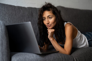 Confident freelance woman working from home, lying on couch with laptop, smiling and focused on online tasks.