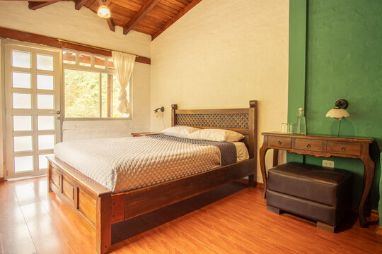 Bright rustic bedroom with wooden furniture, natural light, and mountain view in Ba&ntilde;os, Ecuador. Cozy Andean retreat ideal for relaxation and comfort.