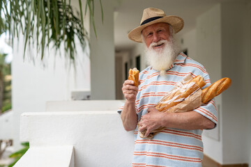 Old man with a long white beard wearing a straw hat and striped shirt, holding fresh baguettes in paper and taking a bite of bread while standing on a balcony with a relaxed expression