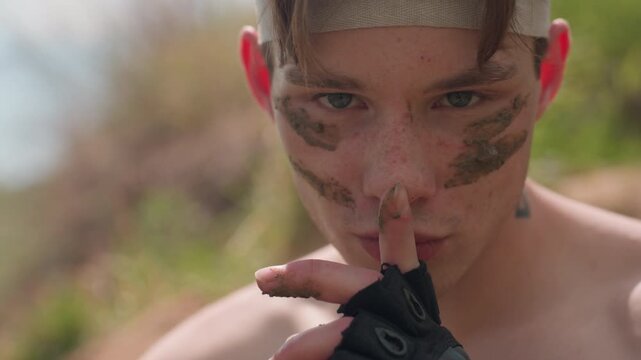 Fierce young warrior with muddy streaks across face stares intensely while placing gloved finger over lips in silent command gesture, standing shirtless in sunlit outdoors, evoking strength