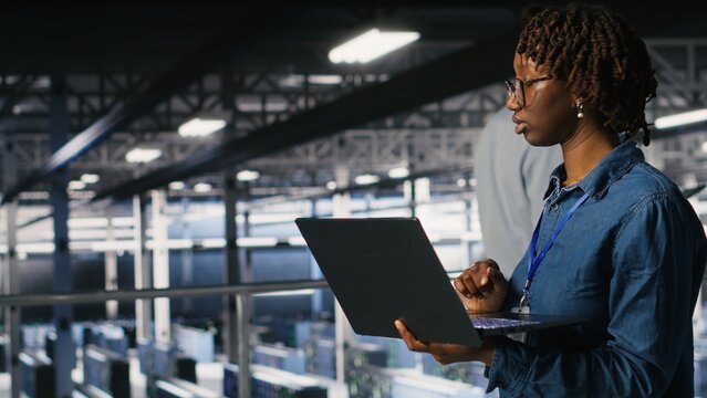 Data center computer scientist doing maintenance tasks on laptop, ensuring optimal performance. African american technician reviewing diagnostics and applying patches to server hub gear