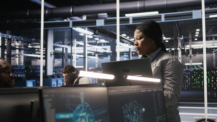 Fototapeta premium Data center manager supervising engineer developing software to analyze neural networks at computer workstation. Team leader and employee observing system performance in AI server room, camera A
