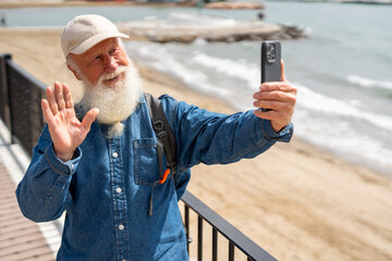 joyful elderly man with a white beard, wearing a denim shirt, beige cap, and backpack, waves while taking a selfie with his smartphone near the seaside on a sunny day