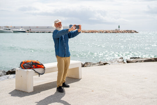 An elderly man with a white beard, wearing a beige cap and denim shirt, takes a photo with his smartphone while standing by the sea, with his orange backpack resting on a white bench - Powered by Adobe