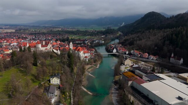 Breathtaking aerial view of Fuessen, Germany, showcasing St. Mang Church, the majestic Hohes Schloss, and the Lech River. The charming medieval town is surrounded by lush greenery and Bavarian Alps. 
