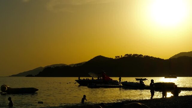 Tourists observing parasailing activity at sunset on beach. Silhouettes of tourists watching a parasailer descend towards the shore as the sun sets behind a hill, casting a warm glow over the scene