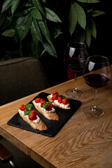 A cozy table is set with two glasses of red wine and a plate of fresh bruschetta topped with tomatoes and basil. Lush plants surround the scene, creating a warm ambiance perfect for an evening meal
